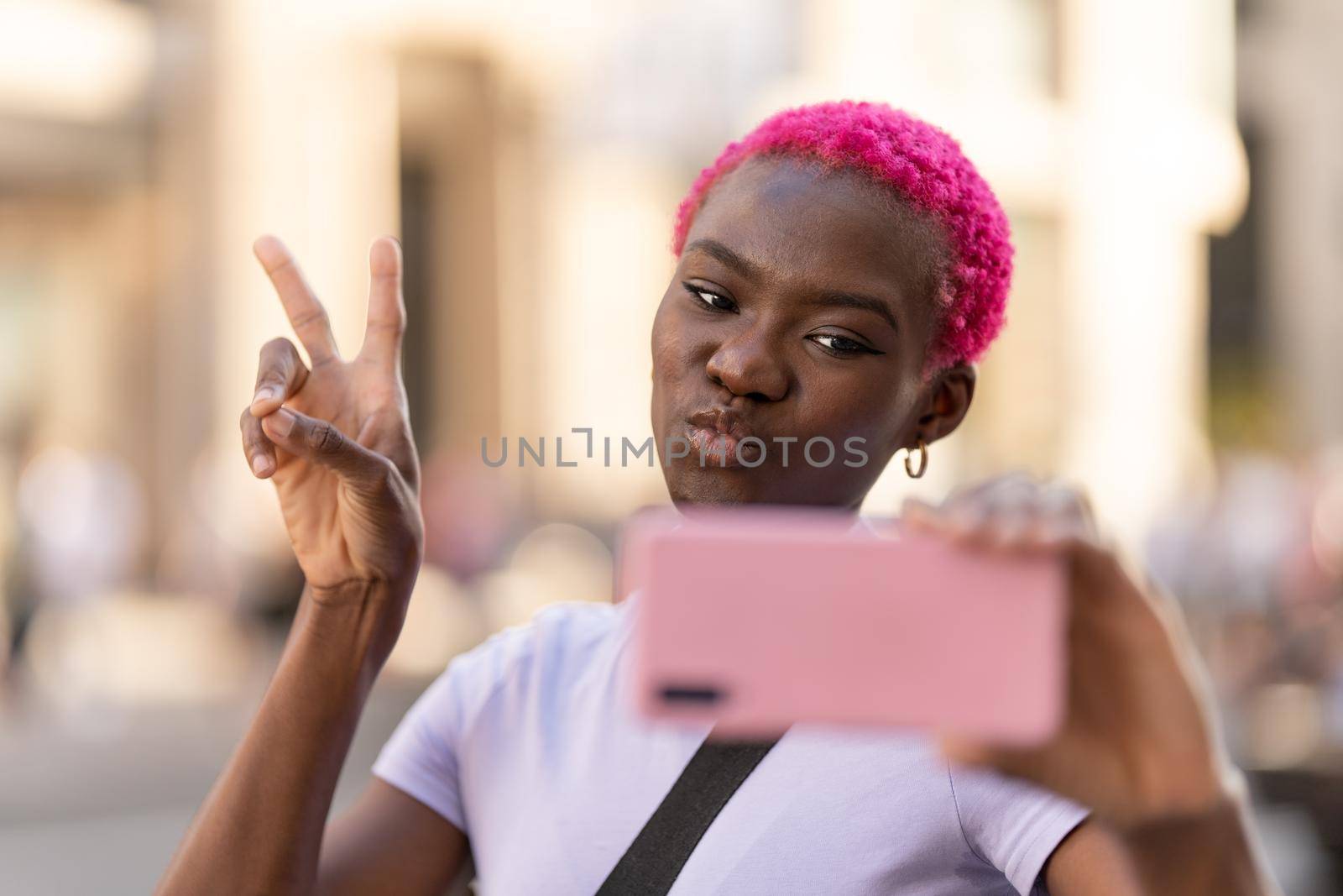 African woman with pink short hair gesturing victory with hand while taking a selfie outdoors