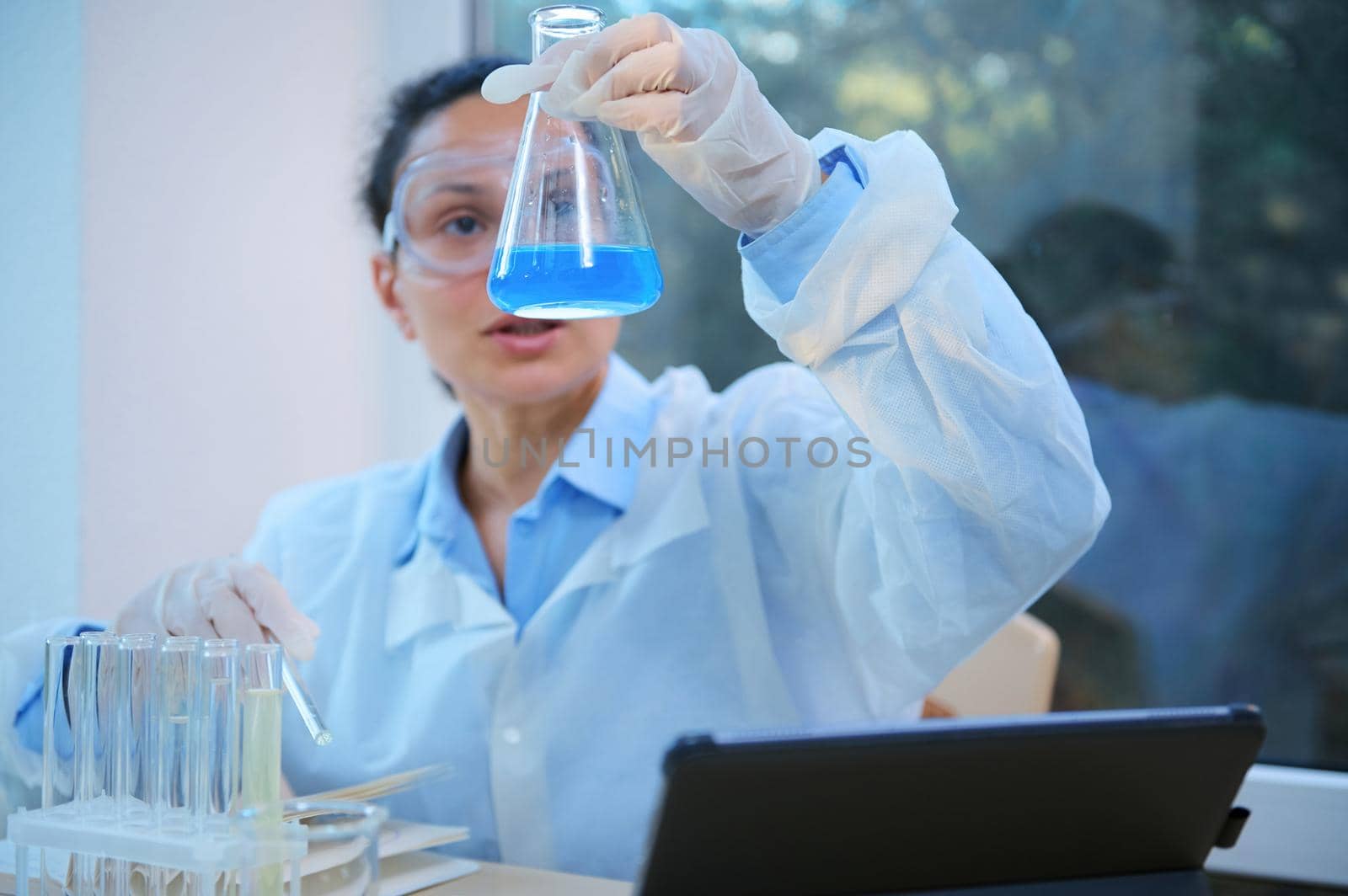 Selective focus on flat-bottomed flask in the hand of woman chemist scientist in white lab coat and protective goggles, inspecting the chemical reaction taking place in blue solution, in laboratory