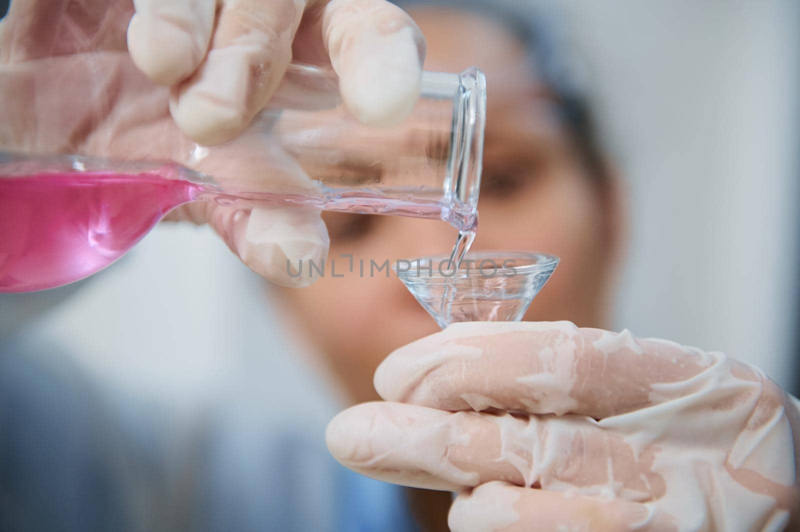Details: Gloved hands of blurred chemist scientist, pouring pink fluid chemical substance from a glass flask through a funnel into a test tube, while conducting experiments in a science laboratory