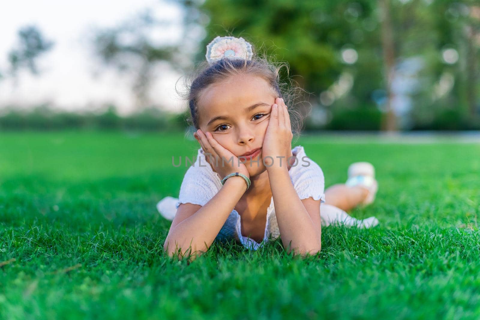 Portrait of a little girl lying on grass with her hands on her cheeks looking at camera.