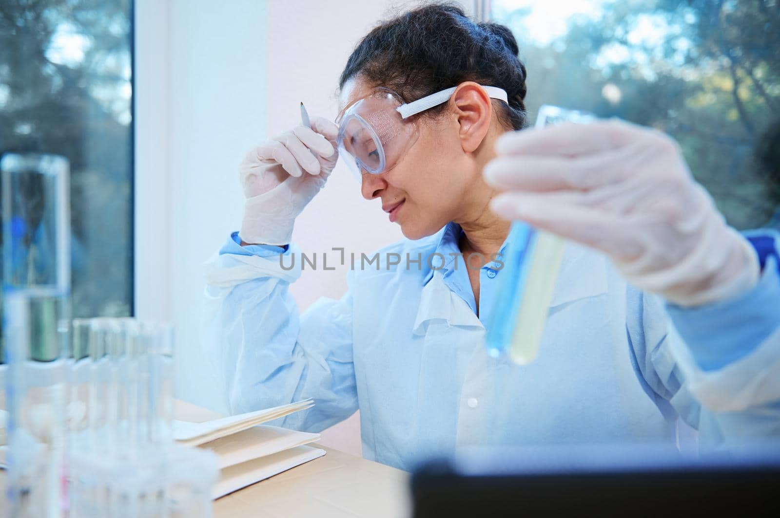 Experienced middle-aged multi-ethnic woman scientist chemist, pharmacologist holds a test tube with blue liquid substance and using tablet for medical research, works in chemical science laboratory.