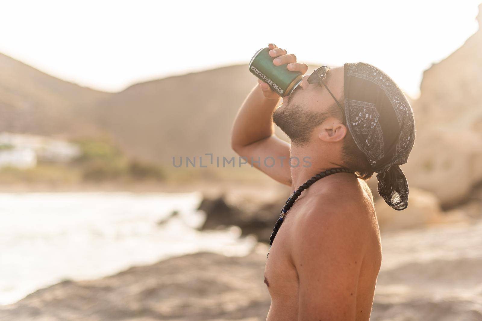 Shirtless man drinking on the shore with light overexposure in the background, sideways