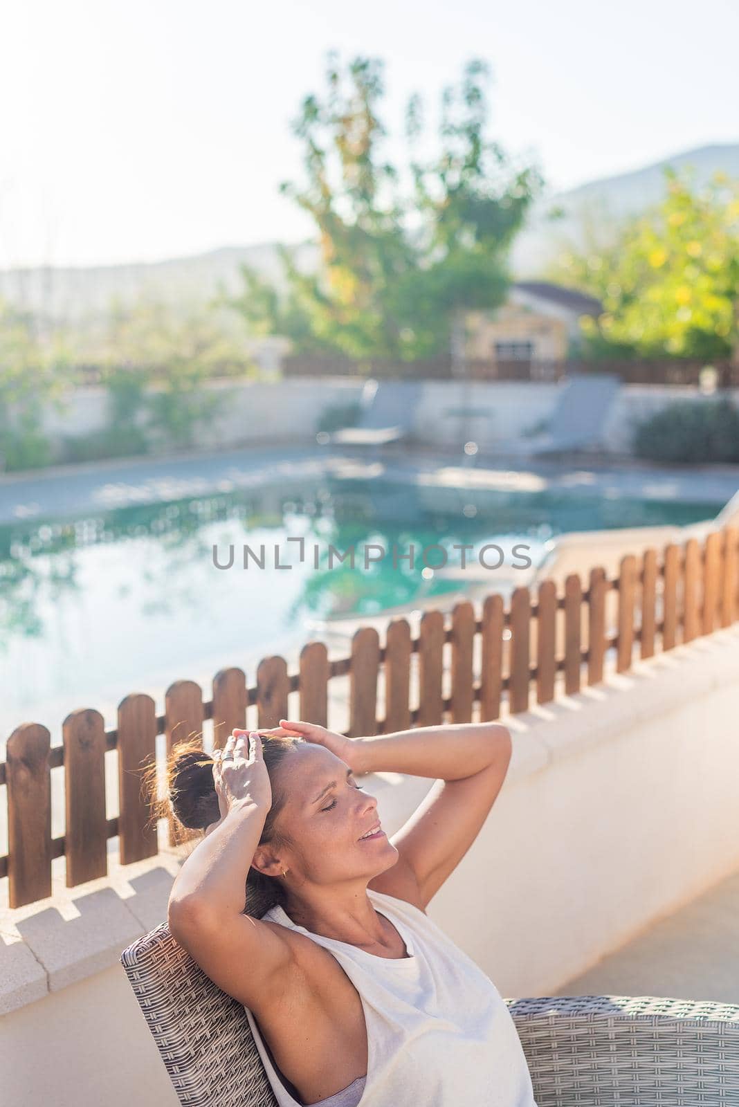 Vertical image of an adult woman with her hands in her head and eyes closed relaxing and enjoying her vacation in a country hotel outdoors sitting near the hotel pool in Almeria, Spain.