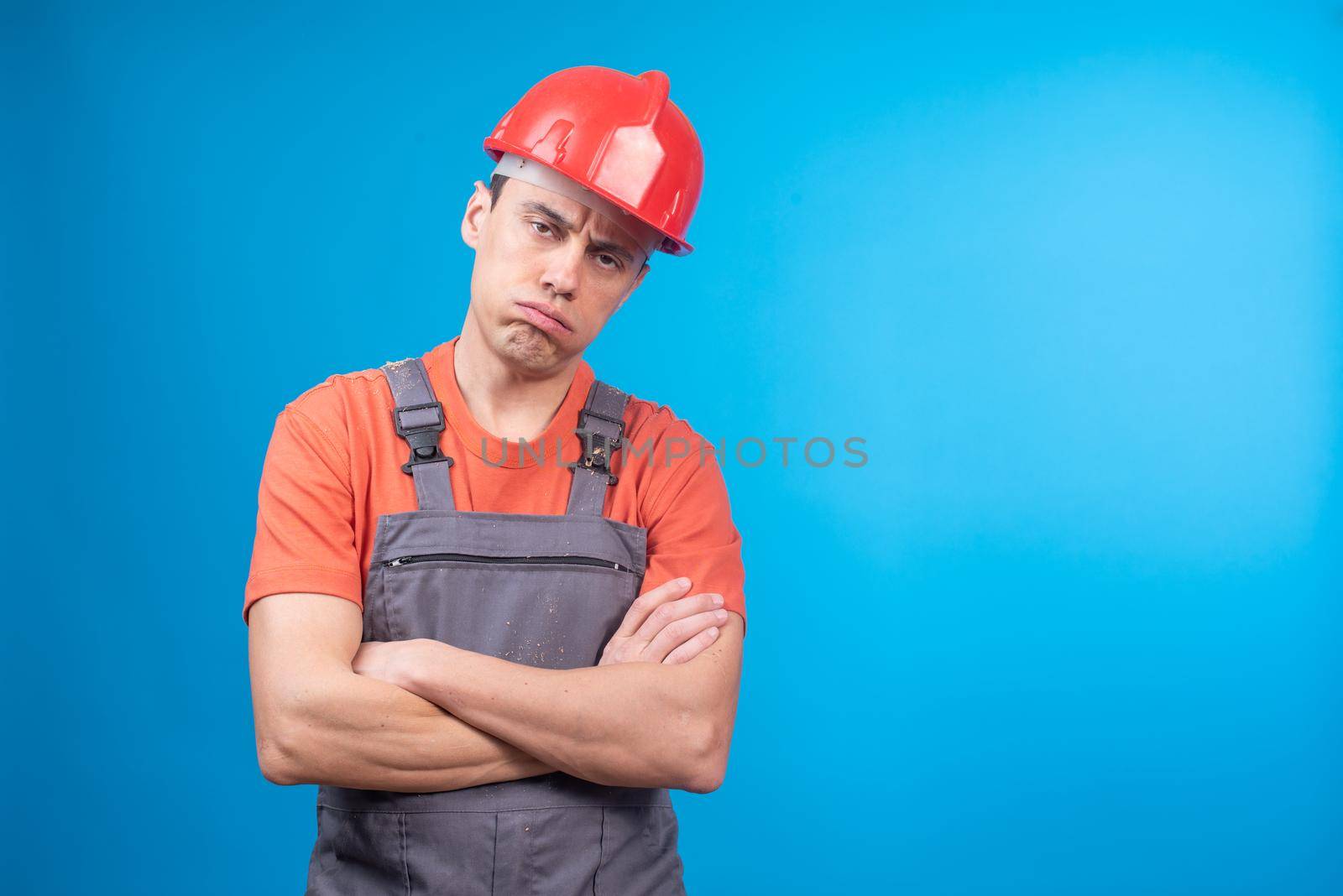 Bored man in builder workwear and hardhat with crossed arms looking at camera and frowning during work against blue background