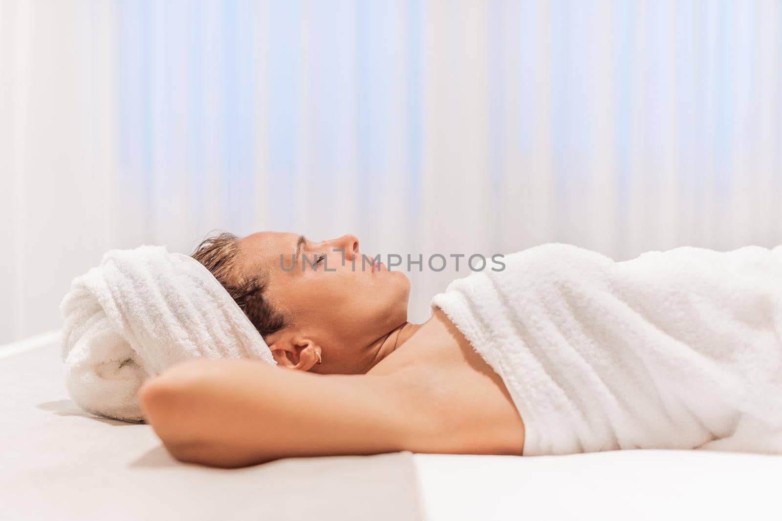 An adult woman wrapped in towels after showering lying in bed with her arms under her head and her eyes closed.
