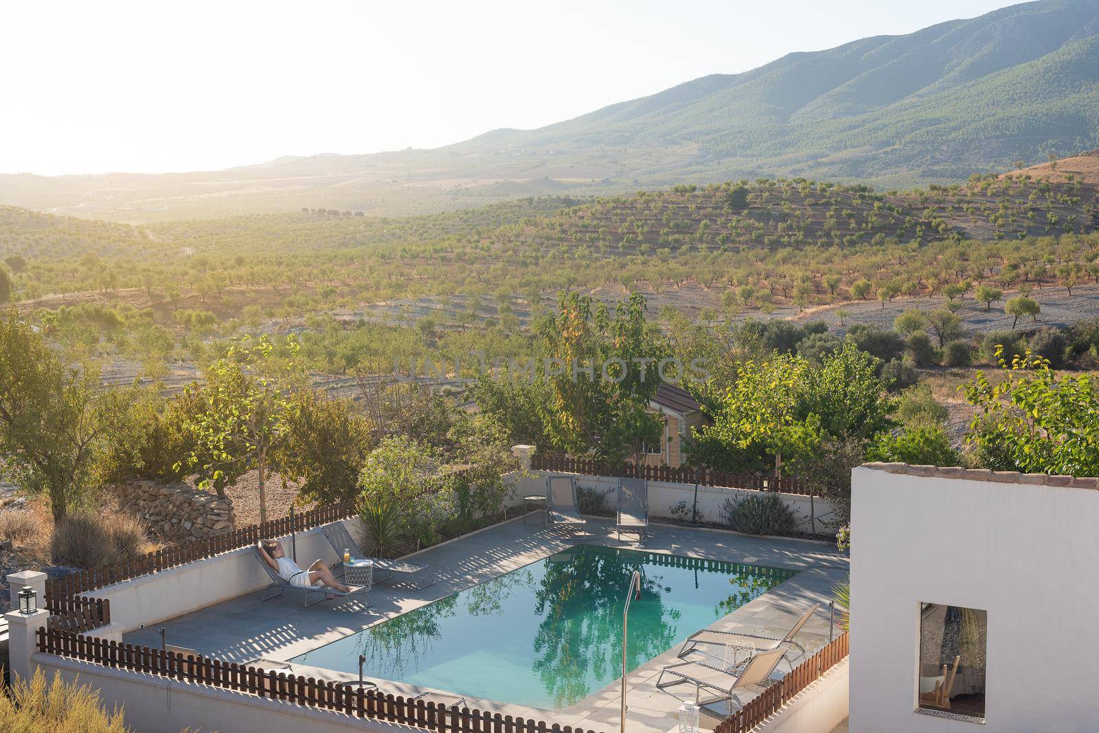 View of a rural house with a swimming pool and a woman relaxing on a deck chair with a landscape of hills and mountains in Almeria, Spain.