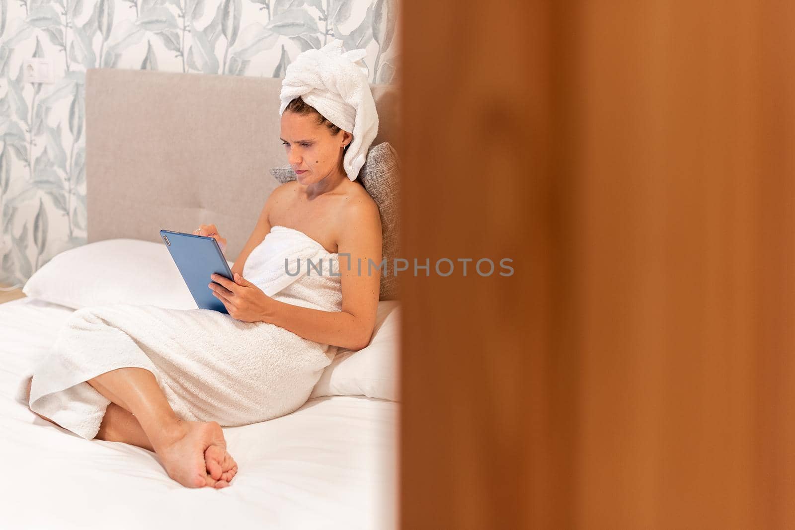 Peeping an adult woman wrapped in towels after showering, lying on the bed using her digital tablet in a hotel bedroom. Concept of hotel.