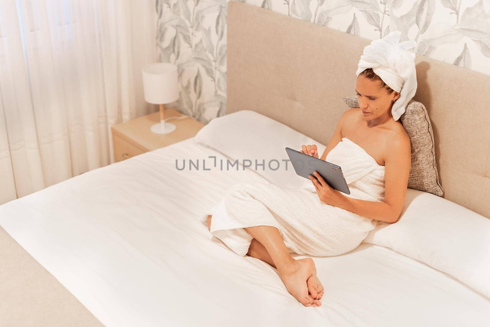 High angle view of a woman wrapped in towels using her digital tablet while lying on the bed of the bedroom of a hotel . Concept of vacation, hotel.