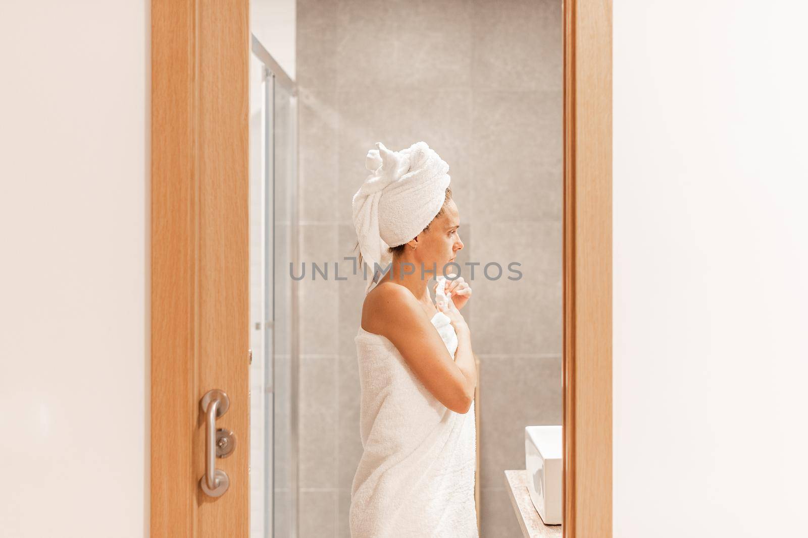 Side view of a caucasian adult woman wrapped with towels after taking shower while looking in the mirror in a hotel bathroom. Concept of hotel.