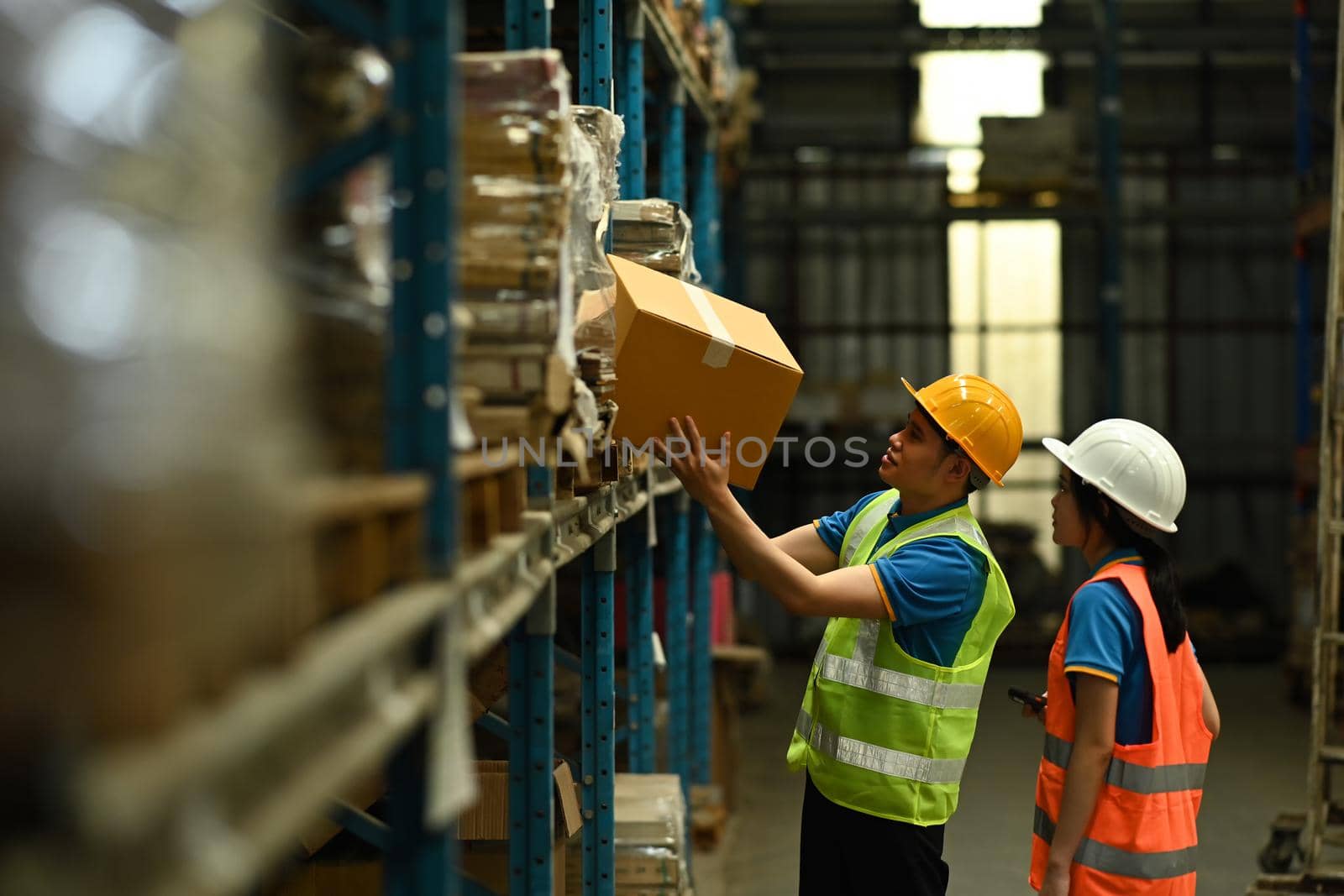 Two storehouse employees wearing hard hats and reflective jackets loading or unloading boxes on the shelf in warehouse.