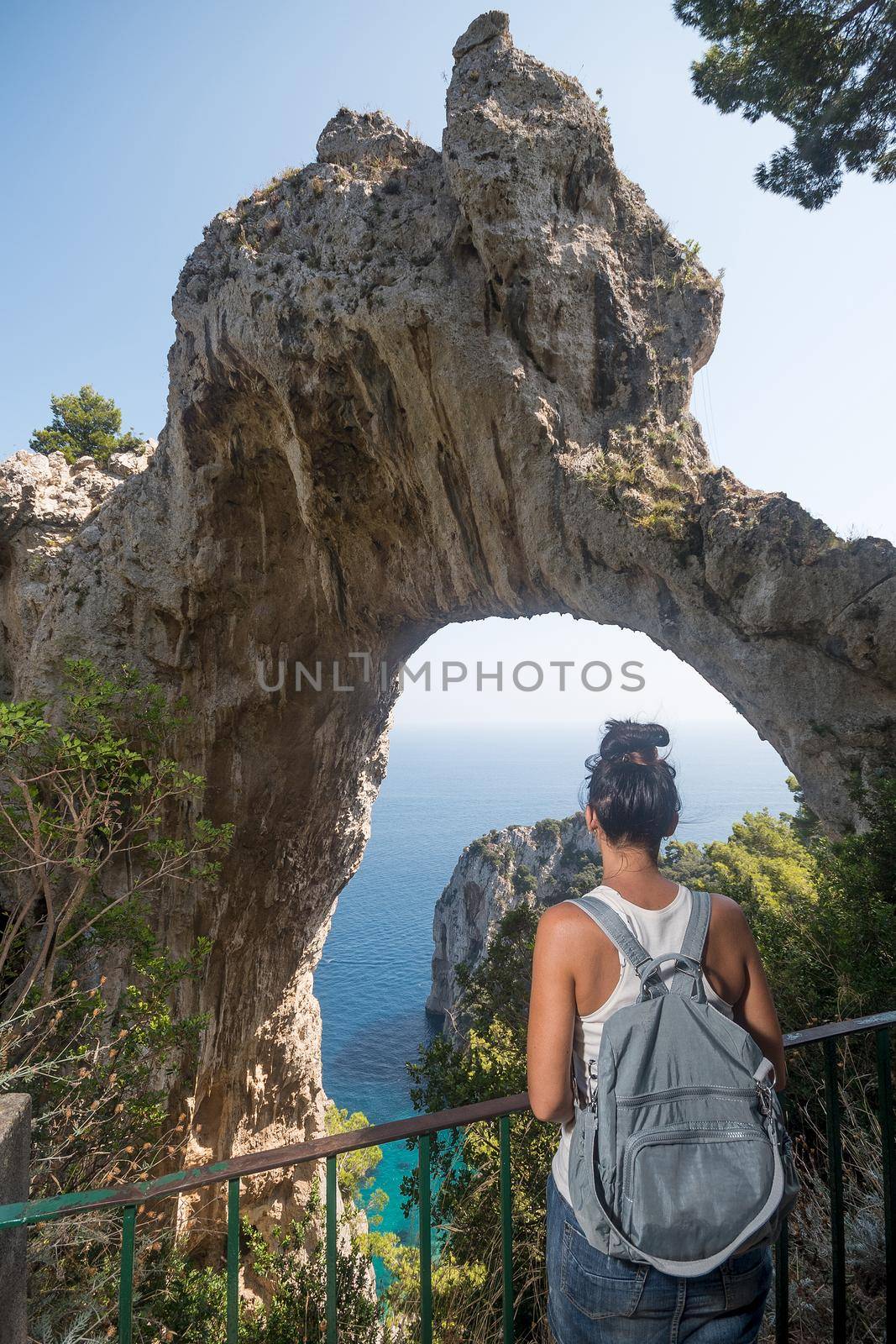 Vertical photo of a woman standing looking to a natural stone formation with the shape of an arch on the sea. Capri, Italy