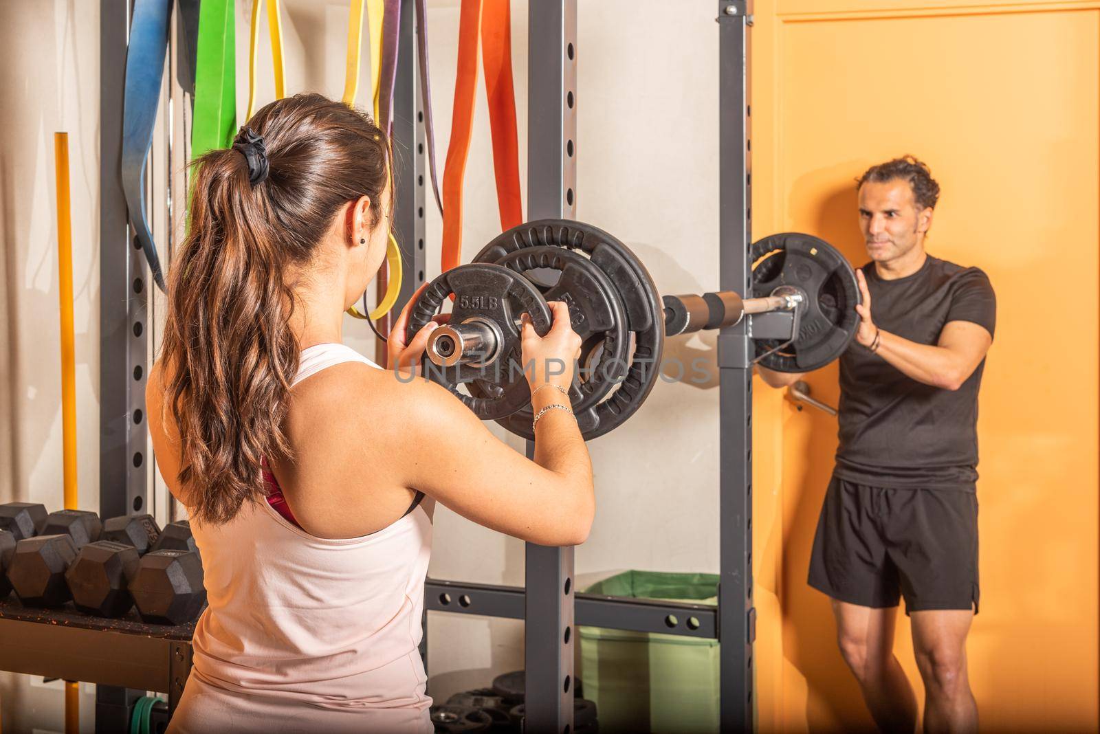 Rear view of woman putting weight plate on bar with help of trainer in gym