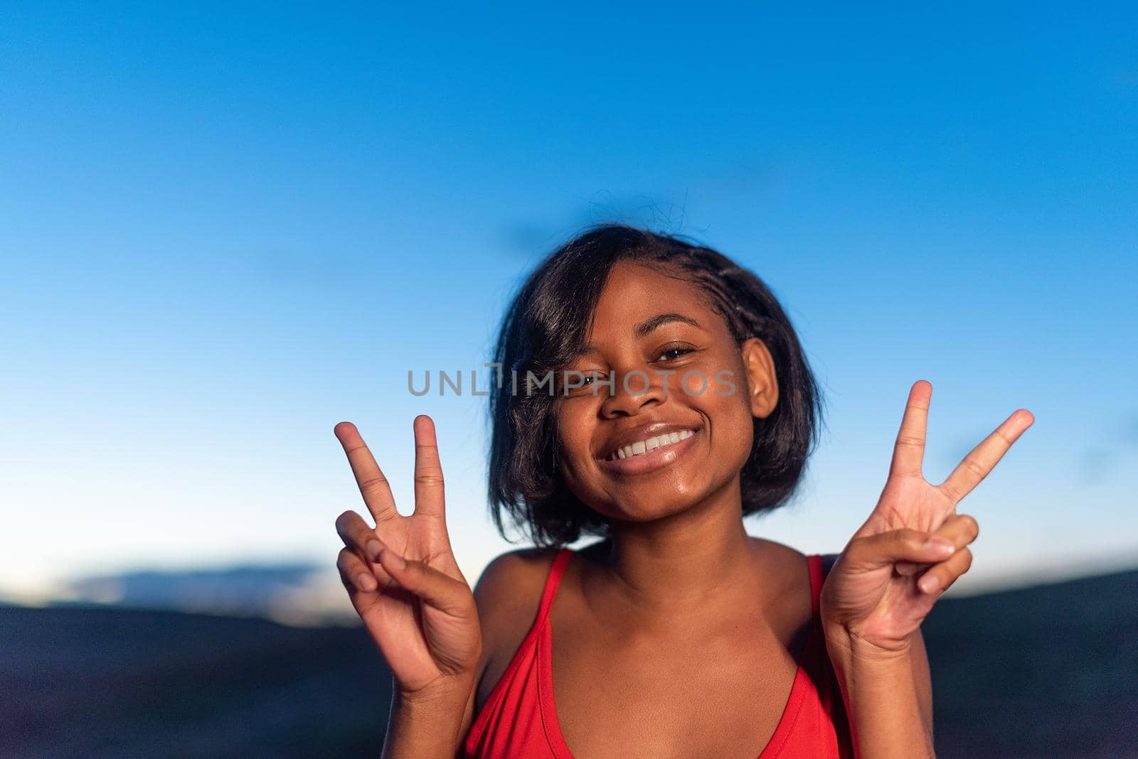 Portrait of a black woman gesturing victory with the hands and smiling to the camera during sunset