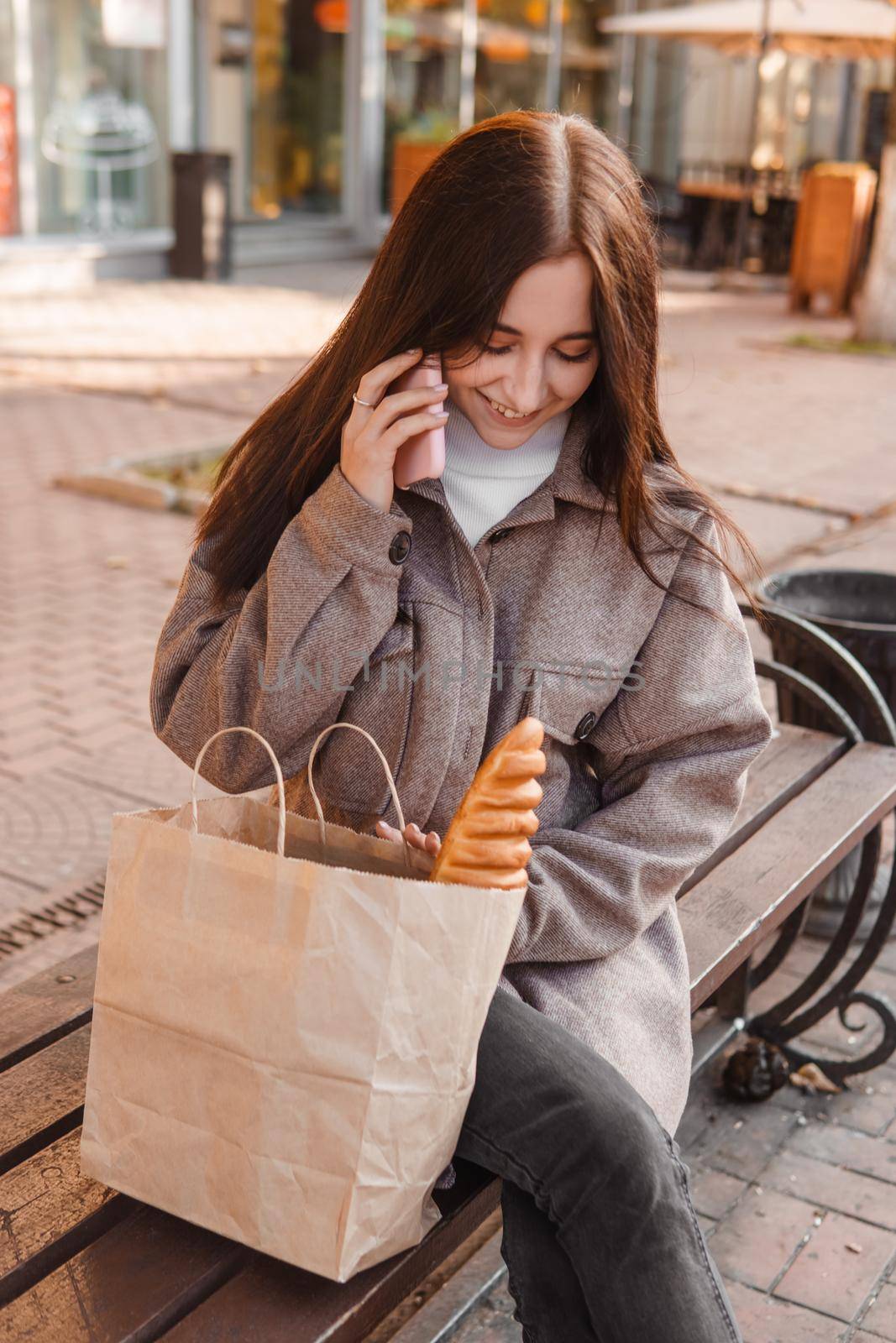 A stylish brunette woman walks around the autumn city. The brunette is sitting on a bench outside with a glass of coffee and a phone, holding a craft bag with a baguette.