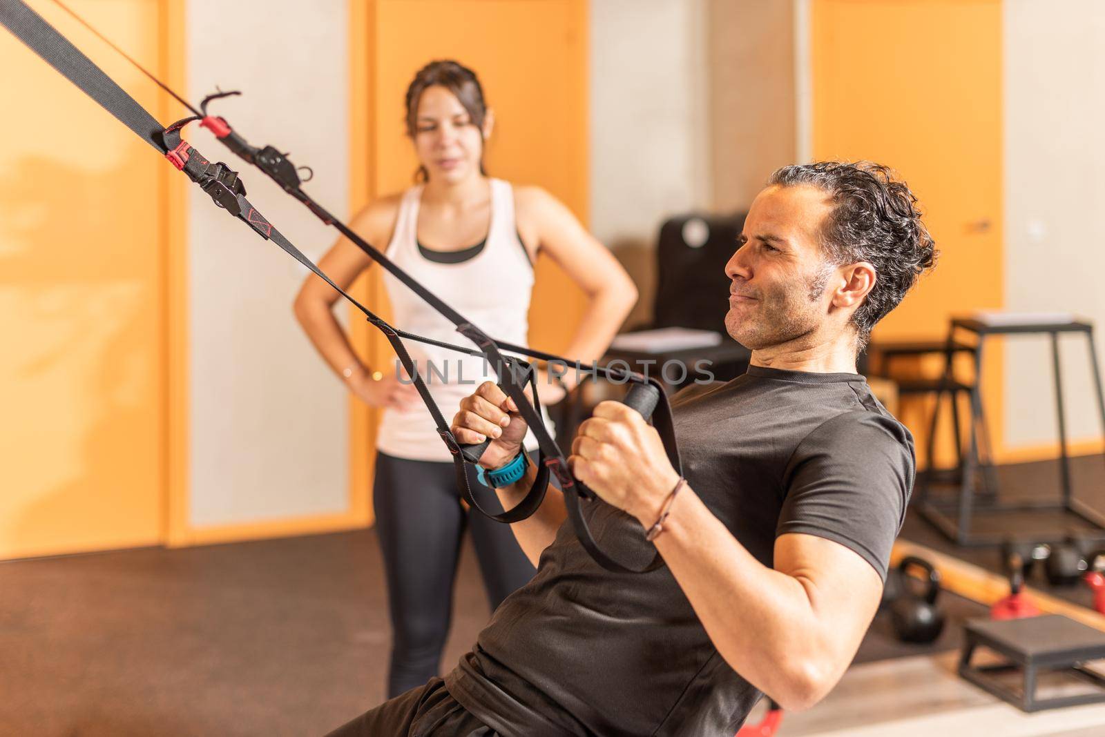 Sportsman doing exercise with trx fitness straps while woman look at him in gym. Concept of trx training.