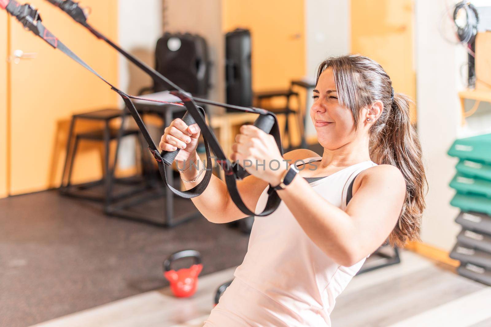 Woman doing exercise with expression of suffering with trx fitness straps at gym by ivanmoreno