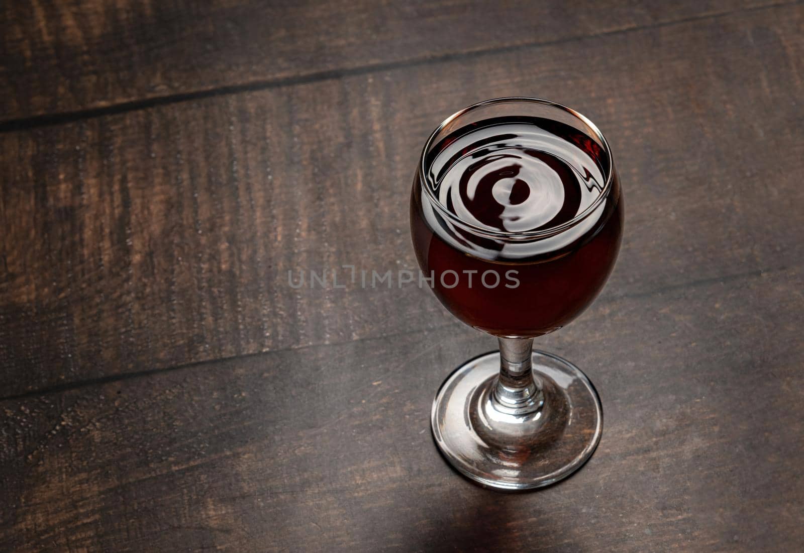 Drops of wine and waves forming in a glass full of red wine standing on a wooden table by Sonat