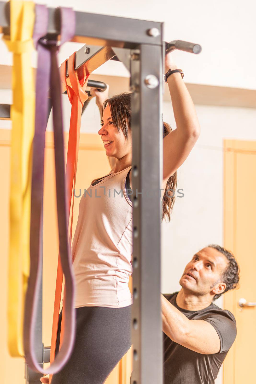 Cropped view of a athlete woman doing pull ups exercise with bar with help of trainer in gym by ivanmoreno