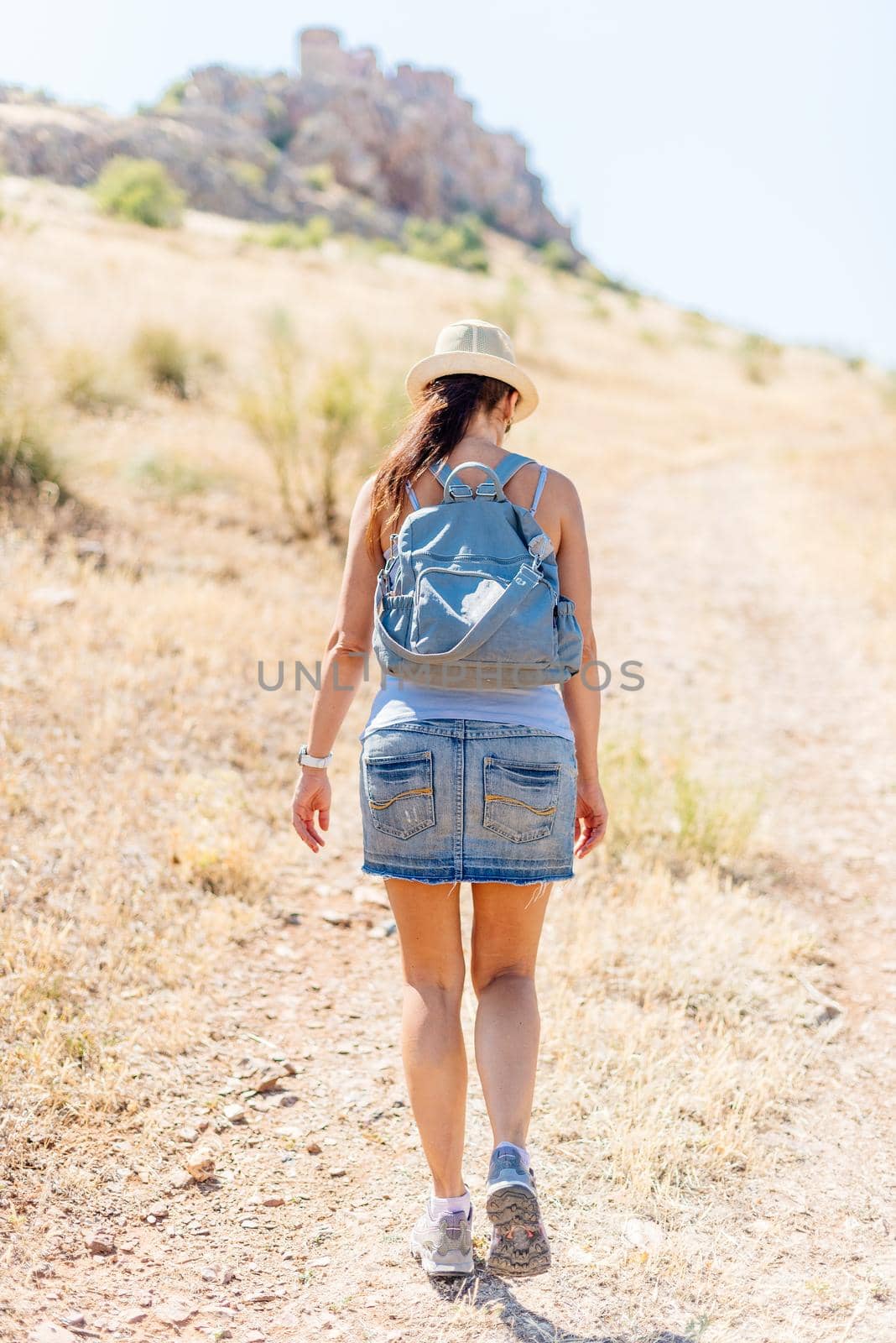 Woman with summer clothes and a backpack walking on a path in a dry grassy field with a castle in the background of the image out of focus. Peñas negras Castle in Toledo Spain