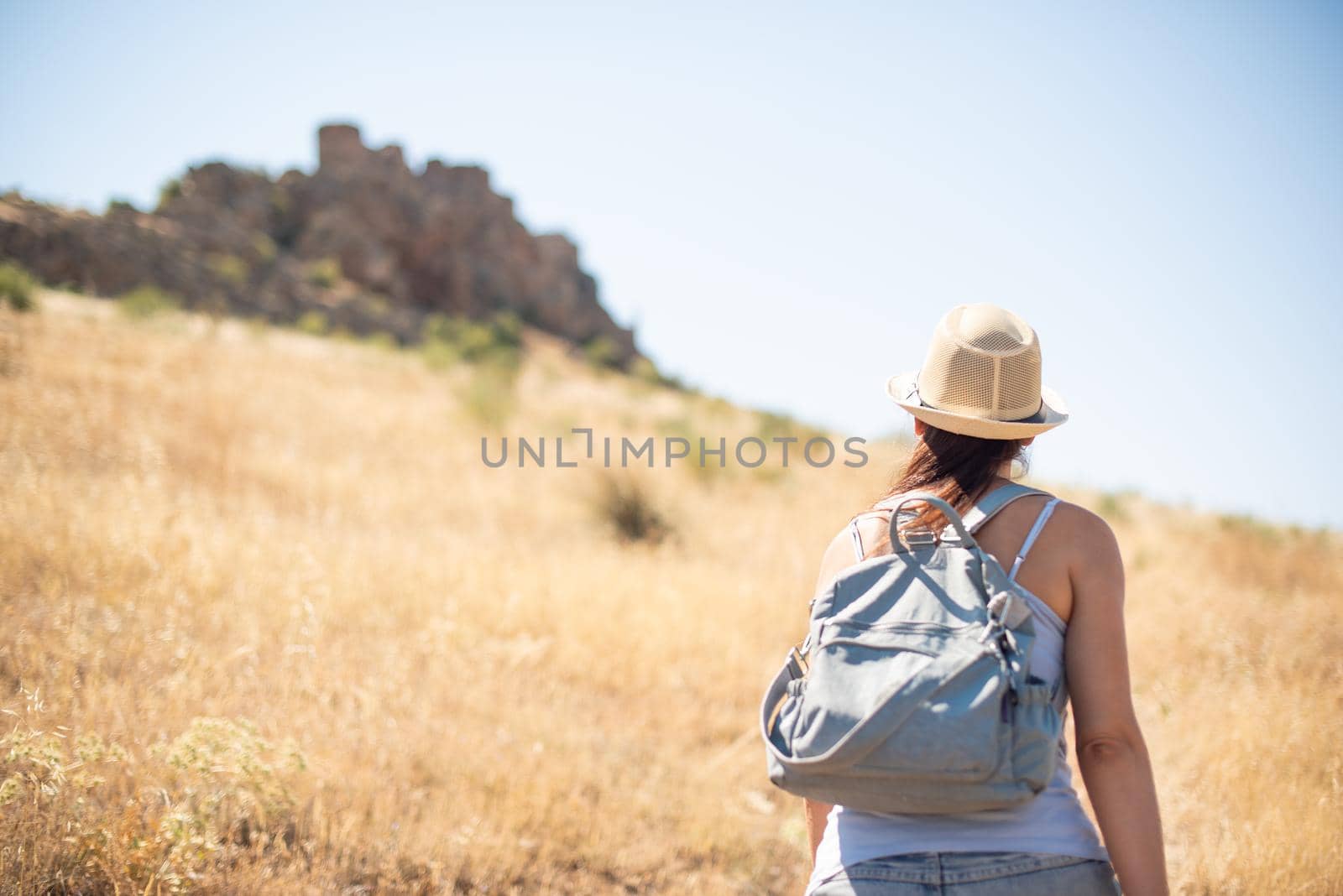 Selective focus on a woman in a hat and summer clothes walking through a dry field towards a Romanesque castle on top of a hill in Toledo, Spain