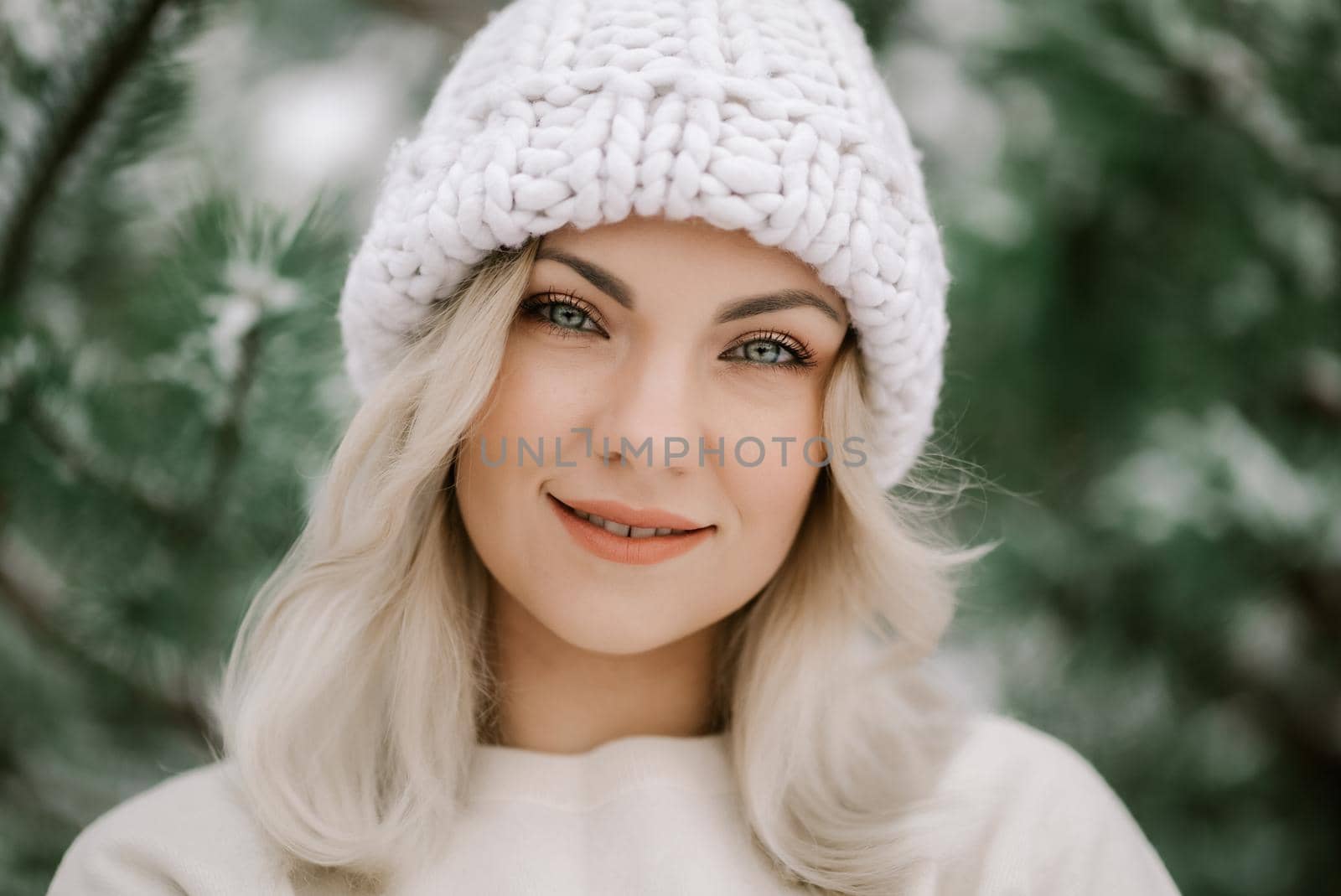 Attractive kind blonde girl smiling, lady in white knitted hat. Portrait of young pretty woman with perfect make-up and hairstyle in forest, winter season. by kristina_kokhanova