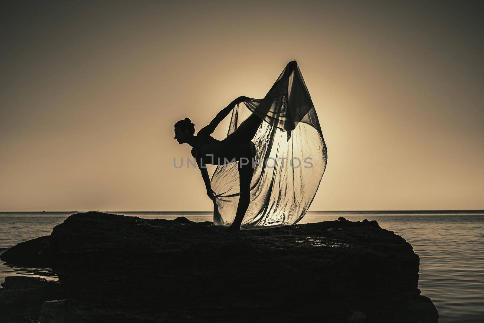 Flexible female gymnast or ballet dancer is dancing with huge silk fabric fluttering in wind on sky and sea background. Concept of tenderness, lightness, art and talent in nature. High quality