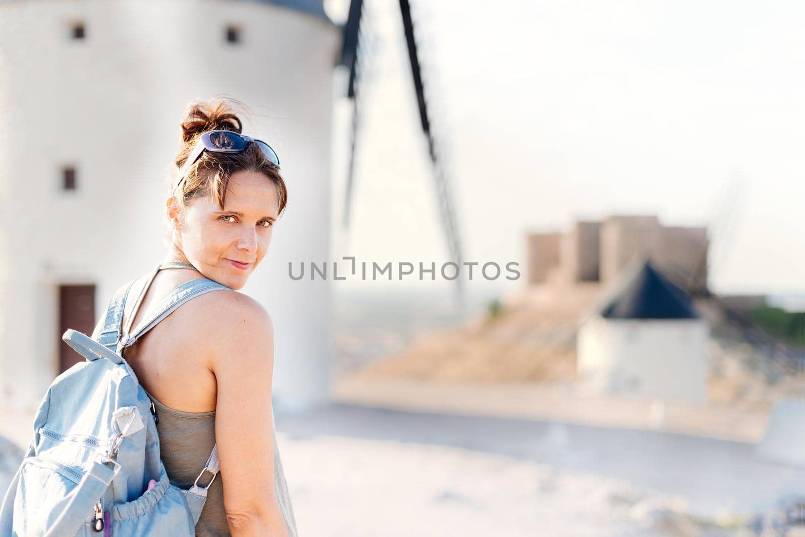 Selective focus on a young woman turning to face the camera standing next to restored ancient windmills and a medieval castle.