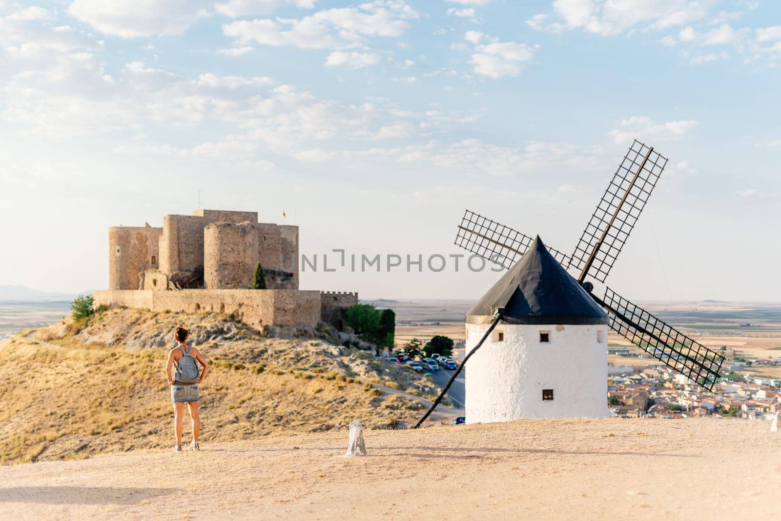 Young woman standing on a hill next to a windmill contemplating a castle on a sunny day in Consuegra, Spain.