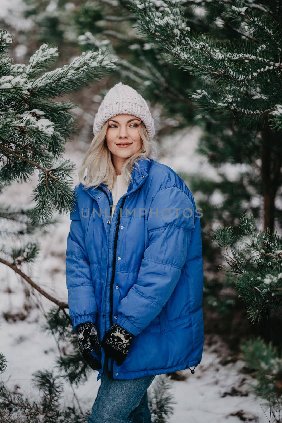 Young beautiful woman posing in forest during winter season. Attractive kind blonde girl smiling, lady in blue coat and white knitted hat. by kristina_kokhanova