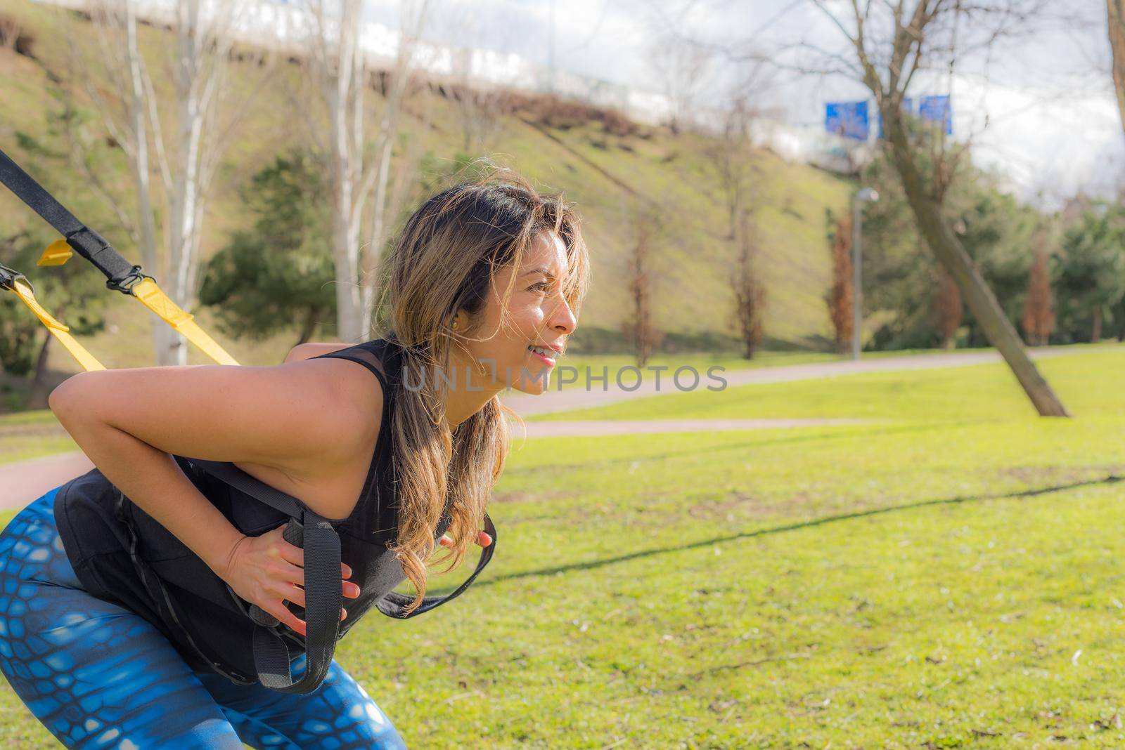 Close up view of an athletic sportswoman doing squatting back exercise with trx fitness straps in park. Adult young woman exercising outdoors.