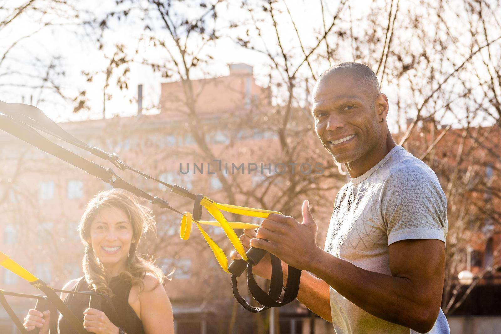 Fitness couple looking at camera exercising with trx fitness straps in the park. Multi-ethnic people exercising outside.