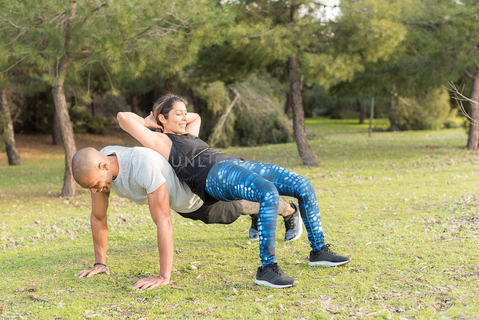 Fitness couple doing back to back lifting exercise in the park. Multi-ethnic people exercising outdoors.