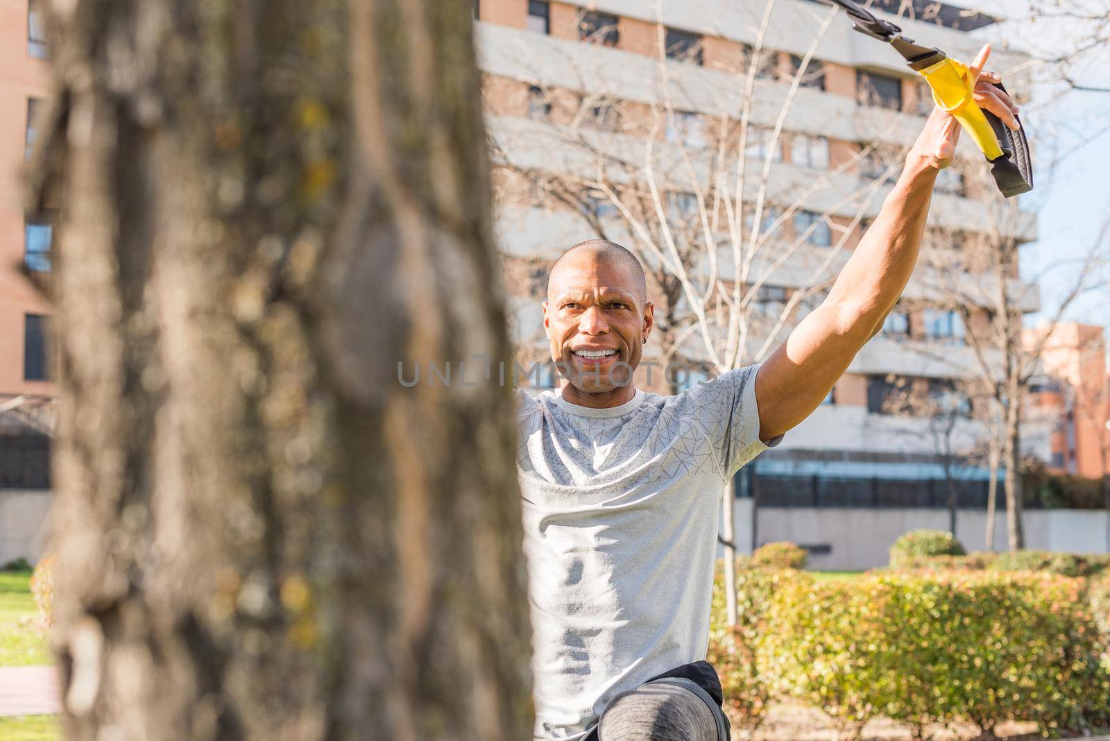 View near tree of a sportsman exercising with trx fitness straps in the park. Adult man exercising outside.