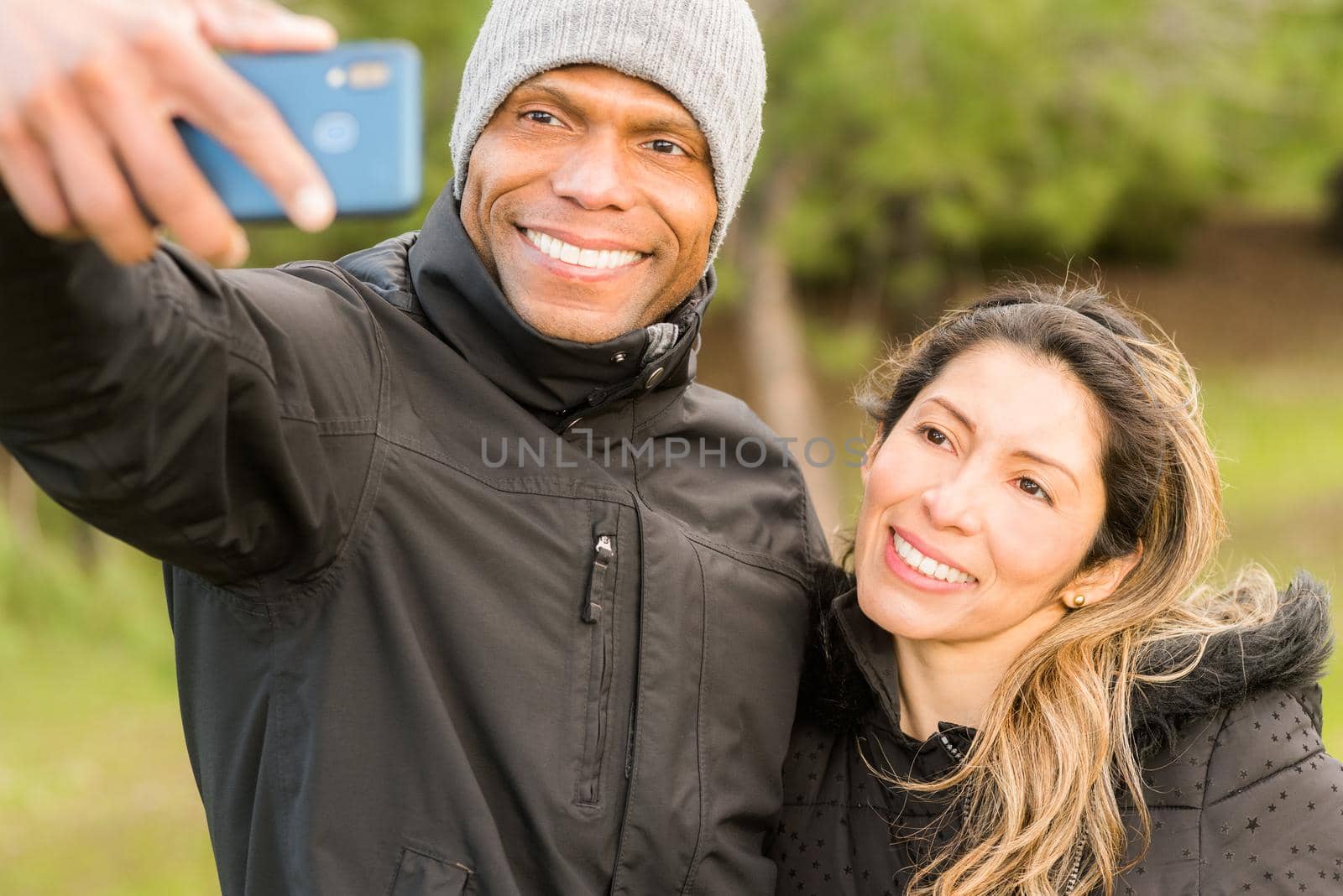 Front view of a fitness couple in warm clothes taking a selfie in the park. Multi-ethnic couple outdoors.