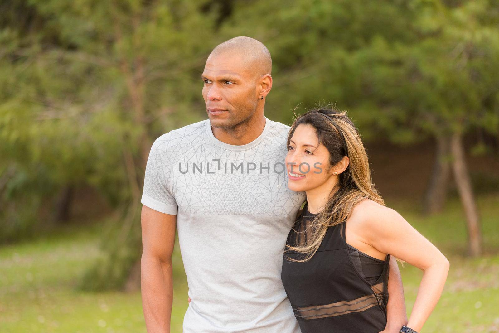 Fitness couple hugging and looking away in the park. Multi-ethnic couple exercising outdoors.