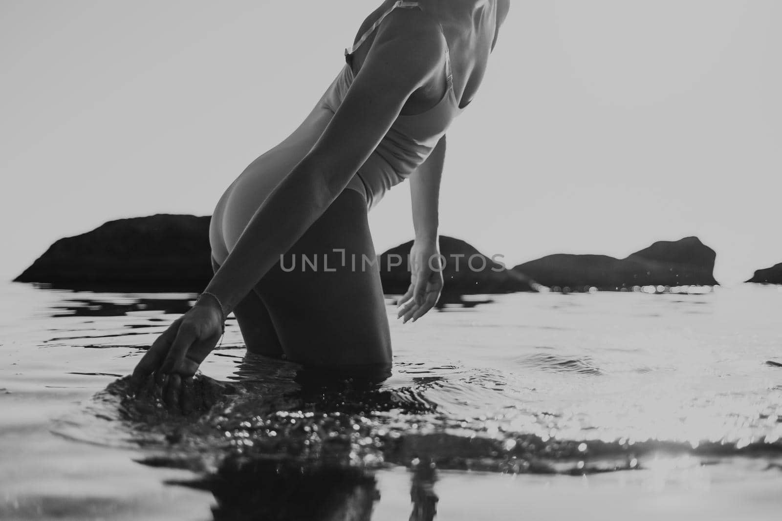 Unrecognizable young sexy woman touching sea water on sunrise background. Girl standing on tropical beach with rocks, lady enjoying summer vacation and ocean. Black and white by kristina_kokhanova