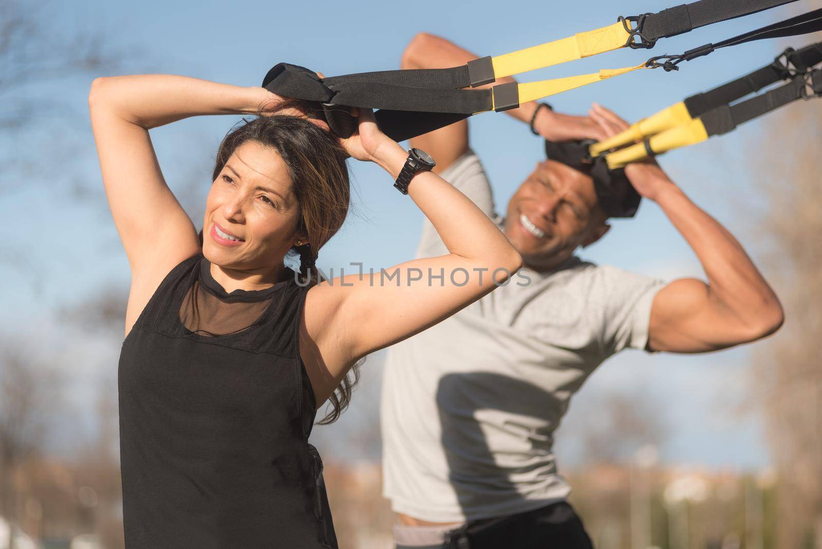 Athletic partners doing biceps and arms exercise with trx fitness straps in park. Multi-ethnic people exercising outdoors.