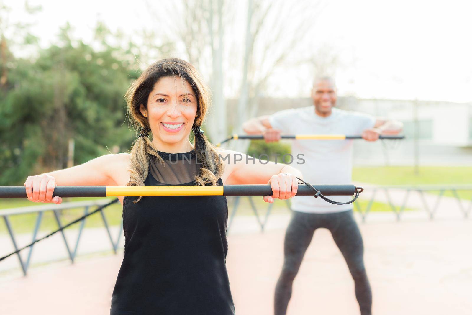 Front view of cheerful fitness couple exercising with an elastic gym stick in the park. Multi-ethnic people exercising outside.