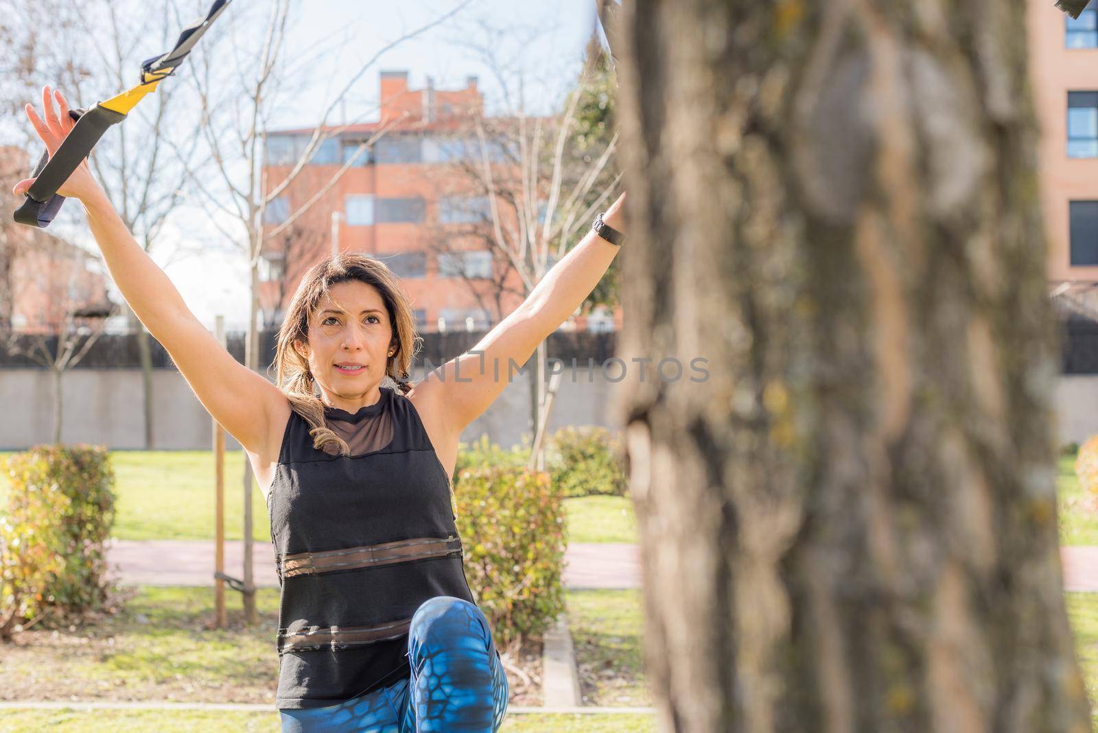 View near tree of a athlete woman exercising with trx fitness straps in the park. Adult woman exercising outside.