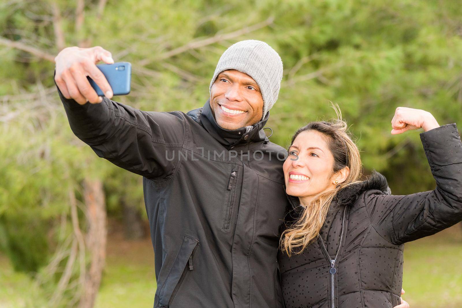 Sportswoman doing gesture flex with one arm while her sporty boyfriend takes a selfie. Multi-ethnic couple outdoors.