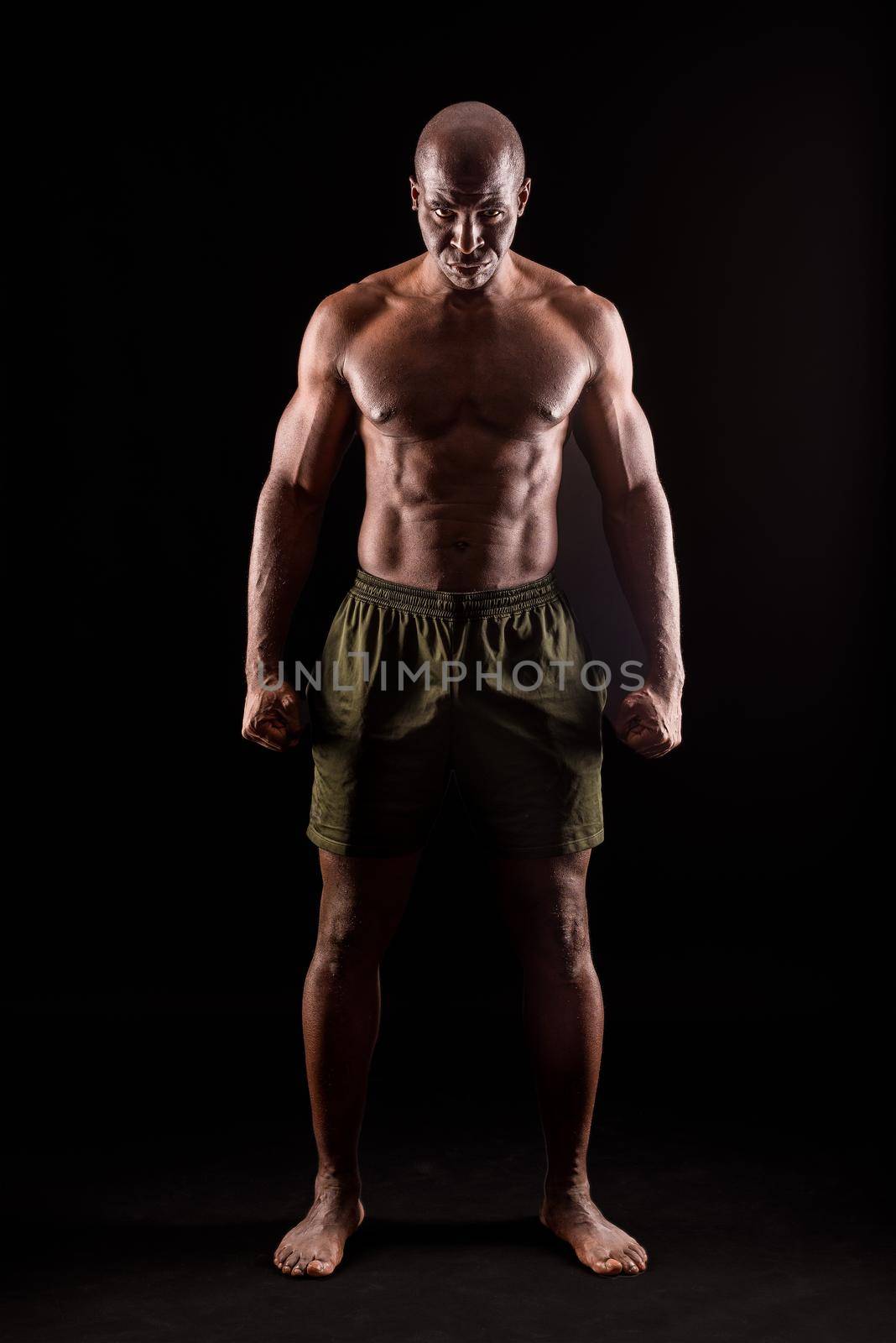 Muscular adult male standing looking at camera with serious expression with arms down and fists clenched. African American adult male standing dressed only in shorts on a black background.