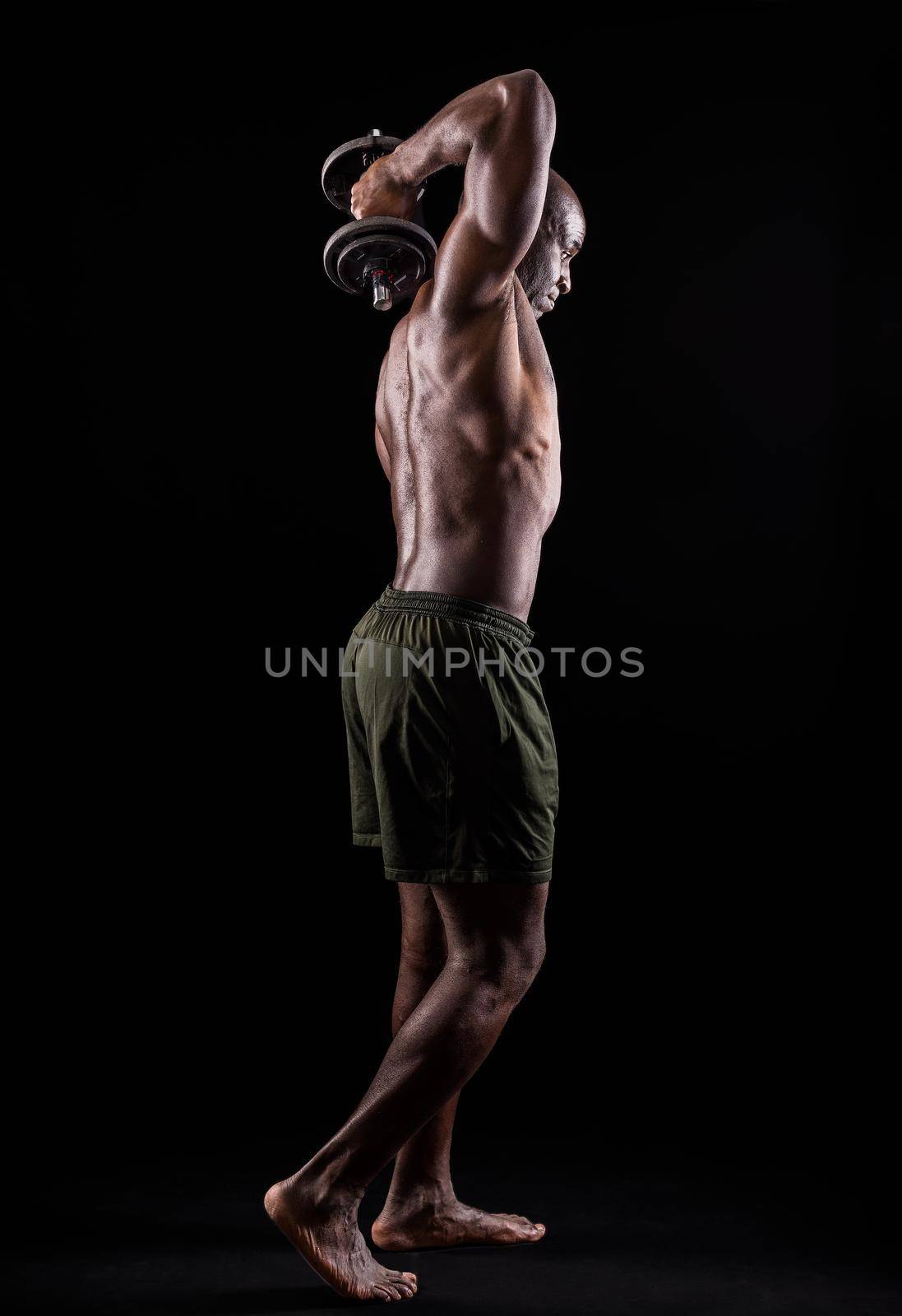 Side back view of a muscular man standing doing triceps exercise with dumbbells with arm raised on a black background. African american adult in shorts doing exercise for arms.