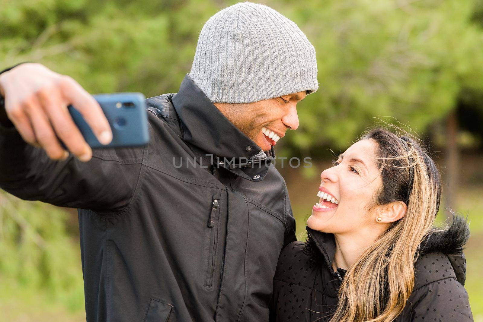 Fitness couple in warm clothes taking a selfie in the park looking at each other. Multi-ethnic couple outdoors.