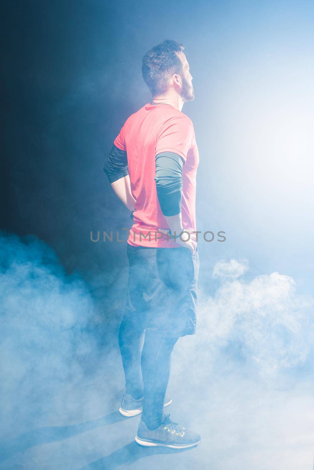 Side back view of an adult male with hands in waist looking forward with smoke around. Caucasian man wearing sportswear in studio with black background.