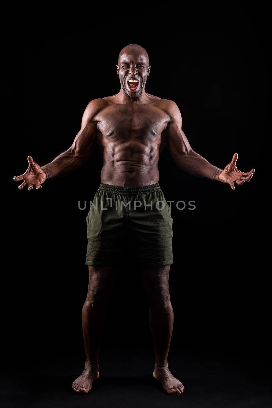 African American man facing front with hands open and shouting at camera. Muscular man with black background standing dressed only in shorts.