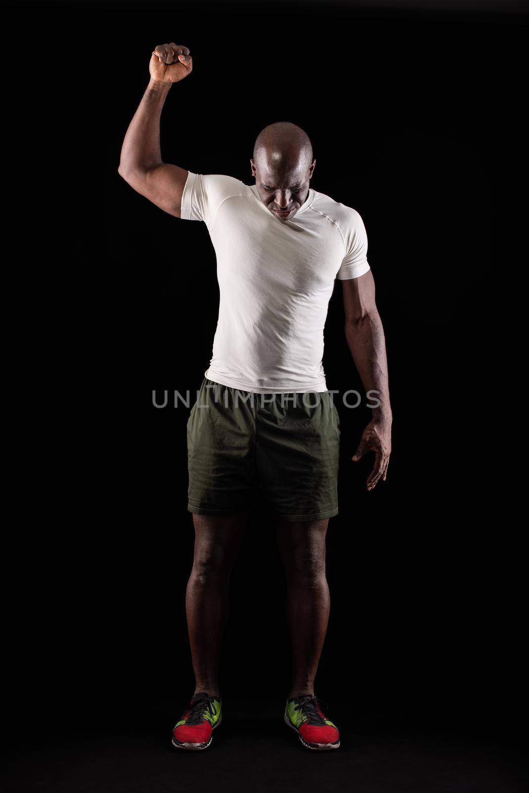 African American male standing with one arm raised and fist clenched while looking down. Muscular adult man in sportswear in a studio with black background.