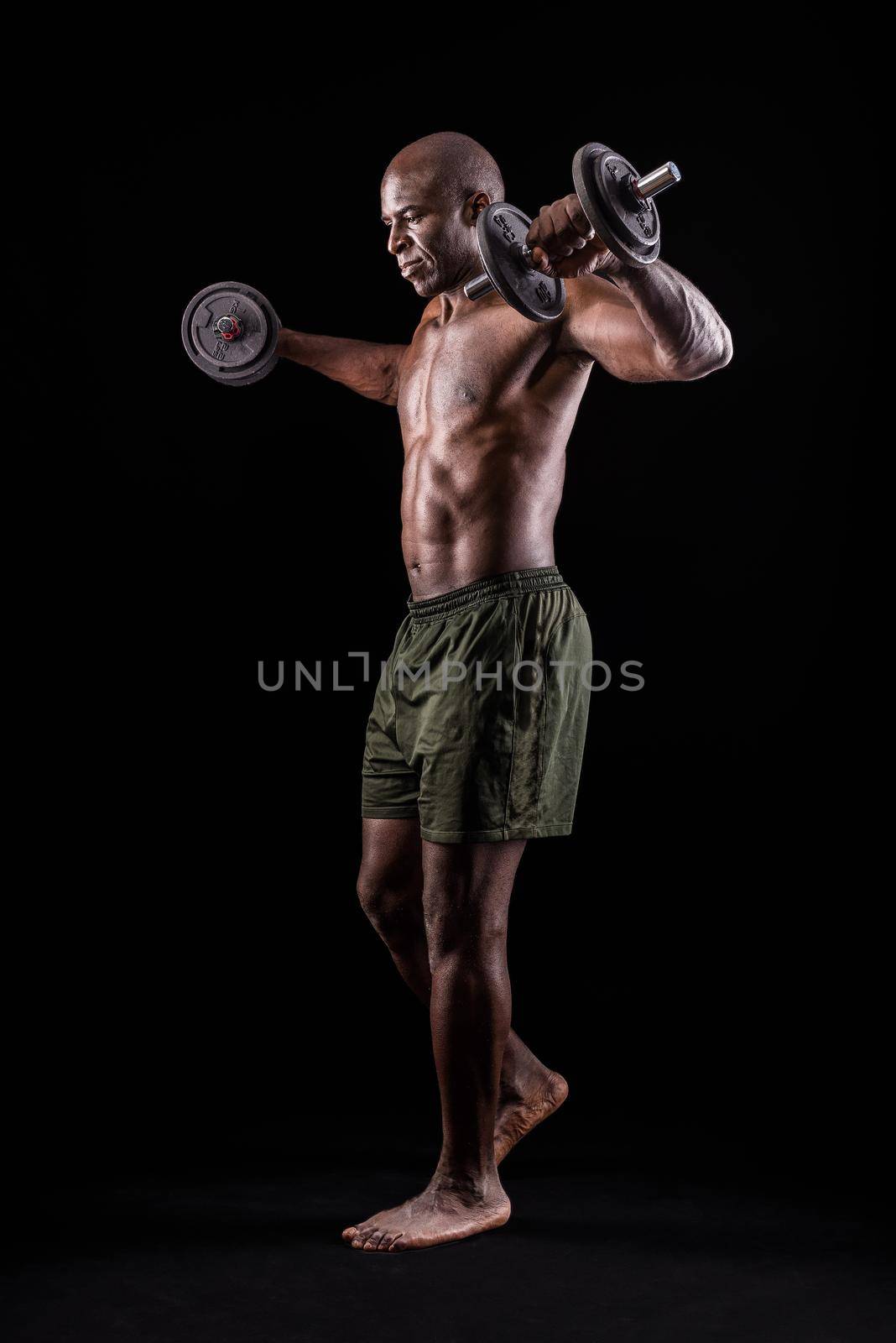 Side view of an athletic adult man doing shoulder exercise with dumbbells on a black background by ivanmoreno