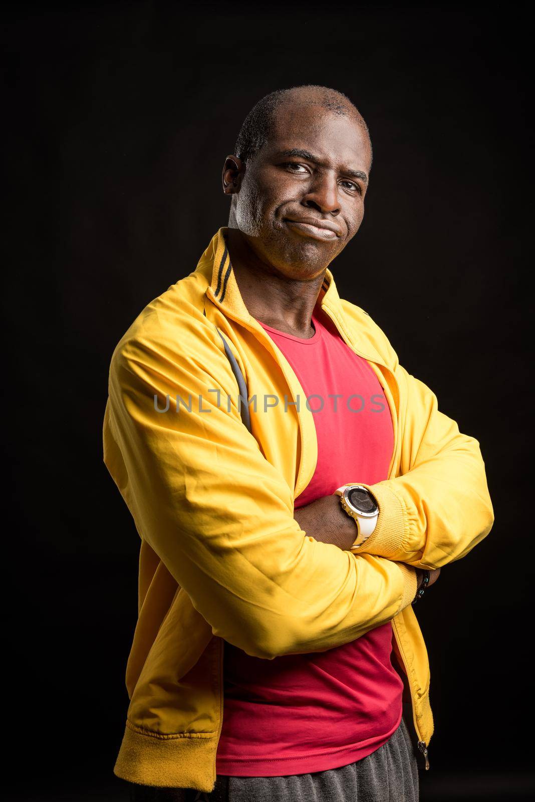 Portrait of an African American man with crossed arms standing looking at the camera. Adult male in yellow jacket and red t-shirt in a studio with black background.