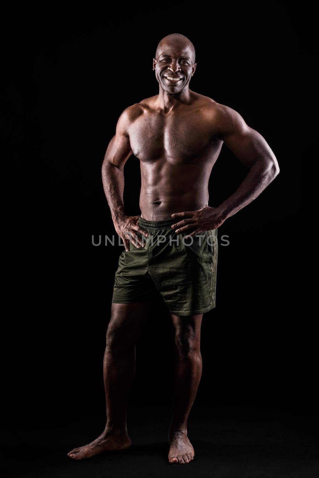 Cheerful adult muscular man standing looking at camera with hands on waist. African American adult male standing dressed only in shorts on a black background.