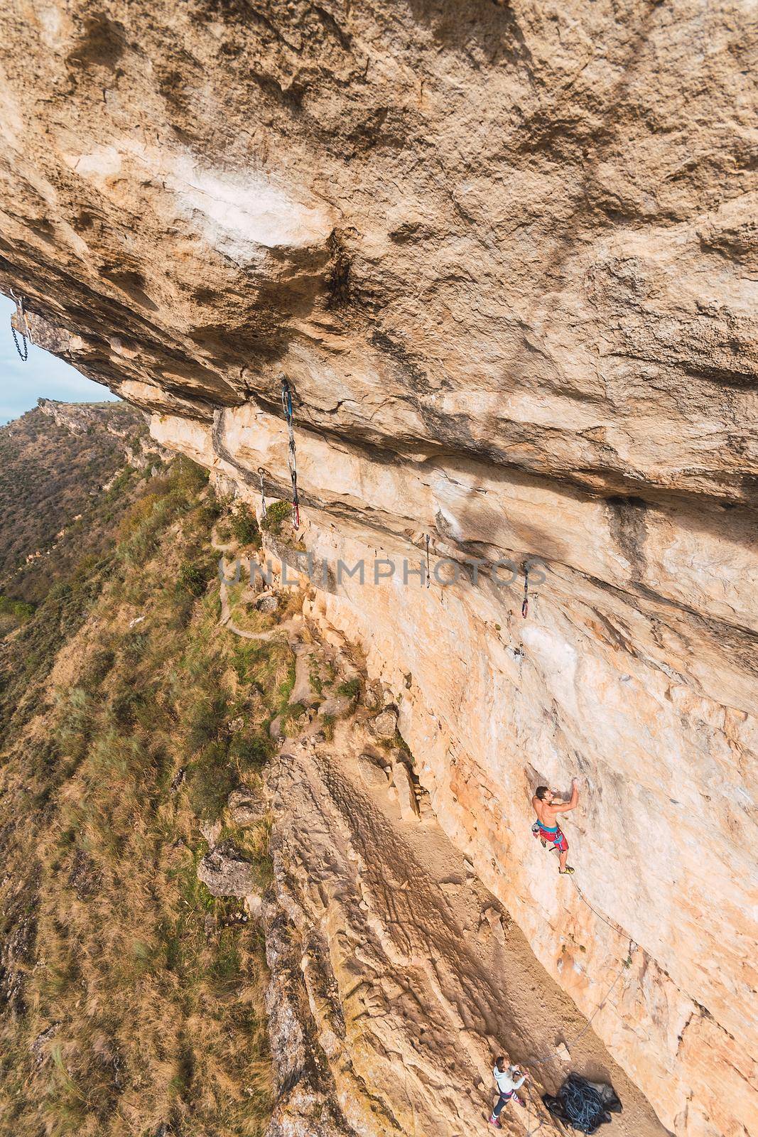 Aerial view of a rock climber climbing a rock formation with people watching from the ground. Vertical view of a climber with bare torso climbing a rock wall.